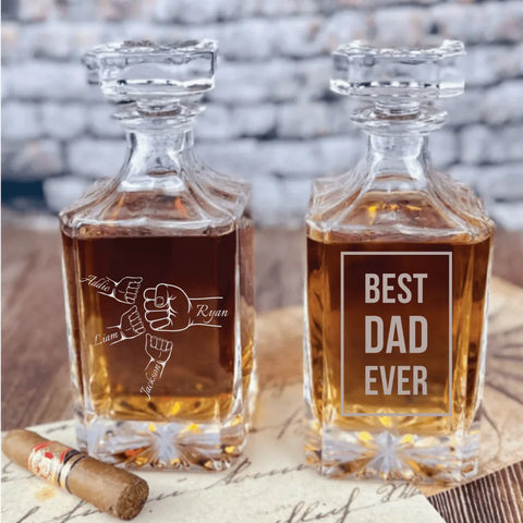Two personalized whiskey decanters engraved for a dad birthday gift, one with a fist bump design and one reading Best Dad Ever, filled with whiskey beside a cigar