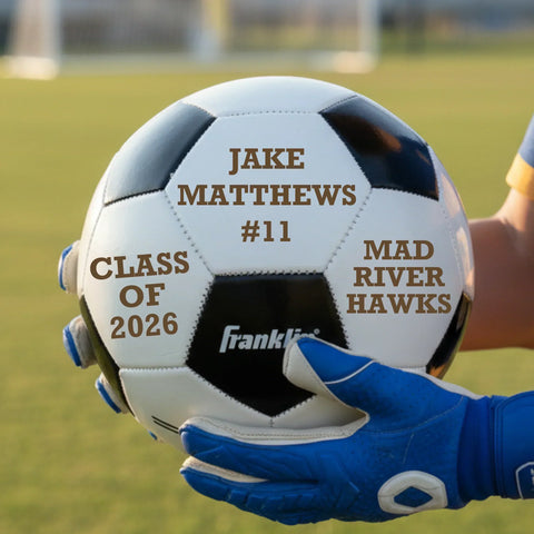White soccer ball engraved with Jake Matthews, number 11, Class of 2026 and Mad River Hawks, held up on a soccer field at sunset