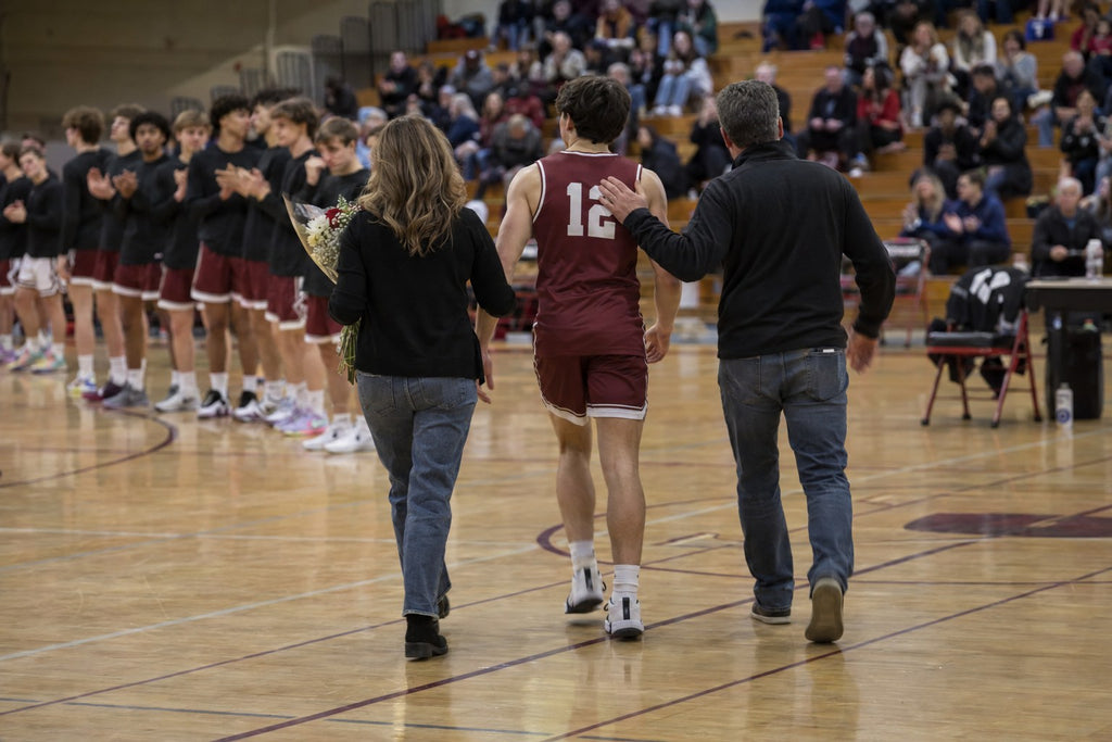 High school basketball senior wearing number 12 walking to center court with his parents on senior night as teammates clap in a crowded gym