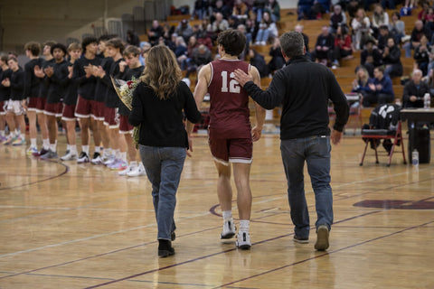 High school basketball senior wearing number 12 walking to center court with his parents on senior night as teammates clap in a crowded gym