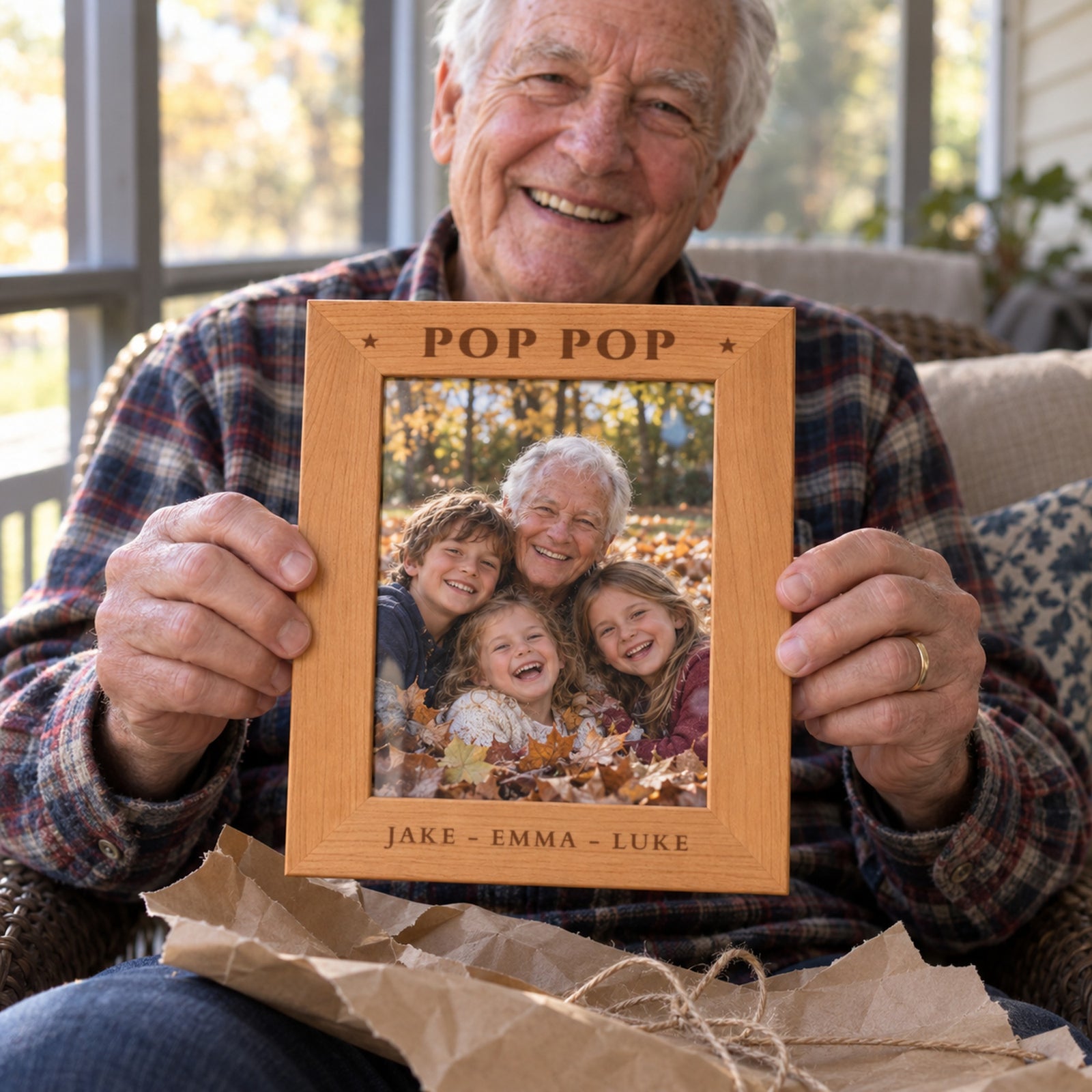 Smiling grandfather on a porch holding up a personalized picture frame engraved POP POP with the grandkids names JAKE - EMMA - LUKE, kraft paper on his lap