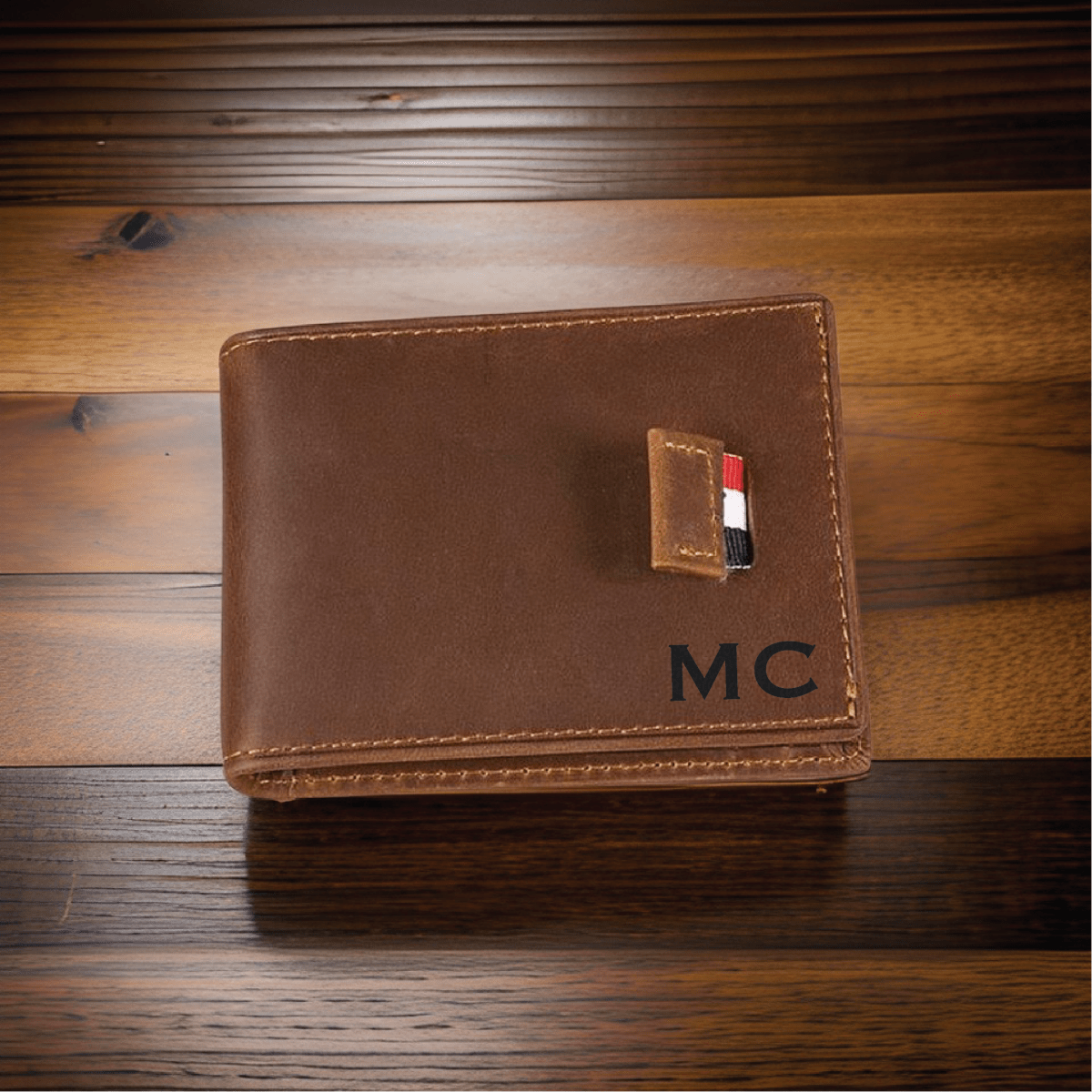 Brown leather bifold wallet with engraved initials and a money clip, photographed closed on a dark wood surface.