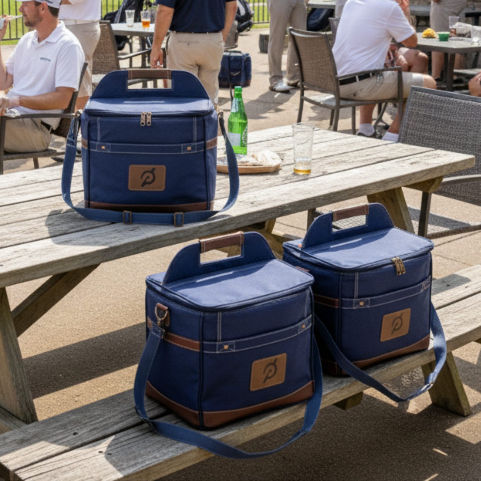 Three navy coolers with tan leather patches and shoulder straps displayed on wooden picnic table at outdoor golf event with beverage bottles nearby