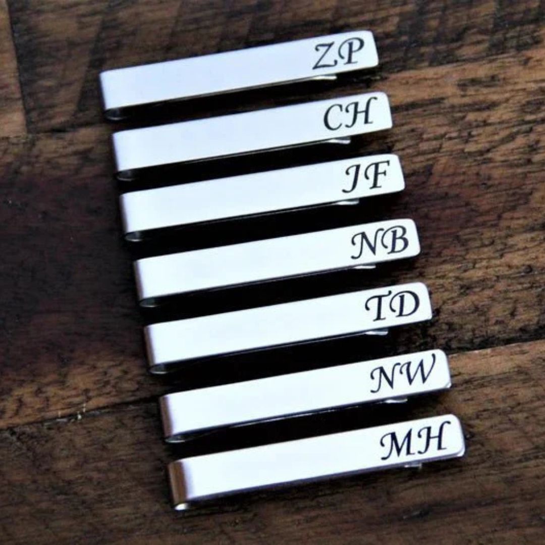 Stack of seven white metal cufflinks with sample monogram engravings including ZP, CH, JF, NB, TD, NW, and MH on dark wood surface