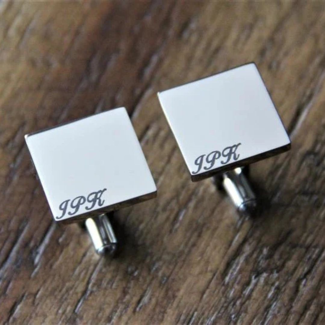 Pair of square silver-toned cufflinks with engraved monogram sample "JPK" on white enamel face, displayed on wooden surface