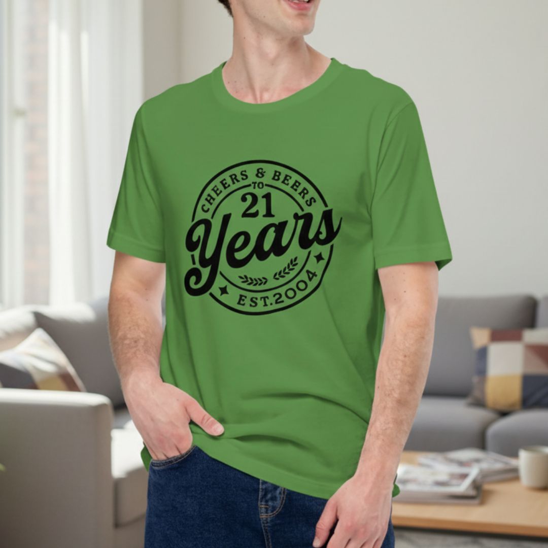 Bright green t-shirt with black circular design reading "Cheers & Beer To 21 Years" and "Est.2003" with decorative stars and leaf flourishes, modeled on male wearer indoors.