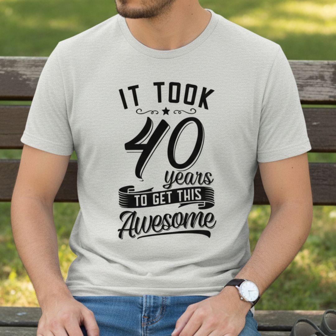 Light gray t-shirt printed with black text reading "It took 40 years to get this awesome" with decorative star and banner elements, worn by man seated outdoors