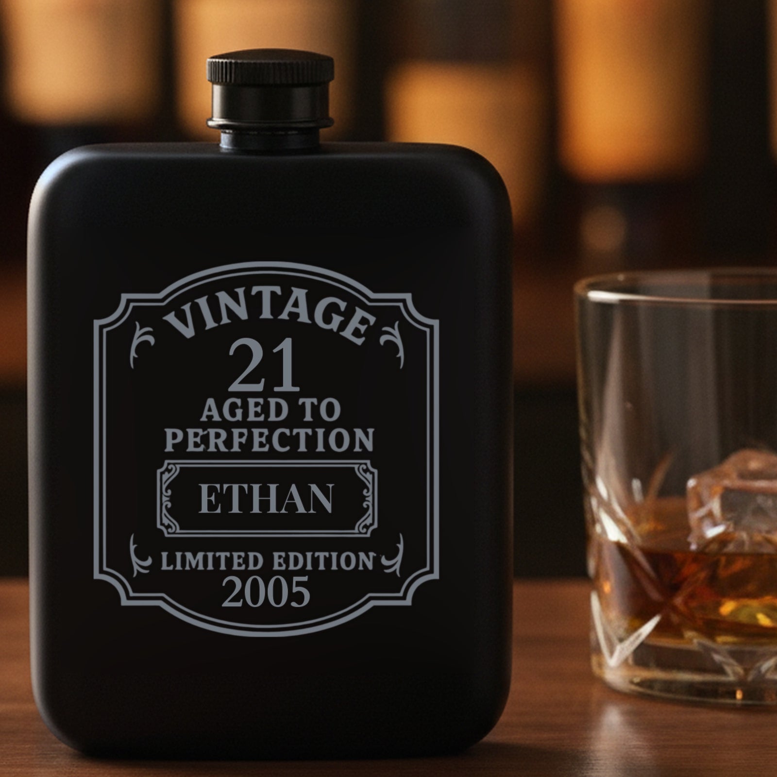 Engraved 21st birthday flask reading Vintage 21 Aged to Perfection, Ethan, 2005, standing on a home bar beside a poured glass of whiskey.