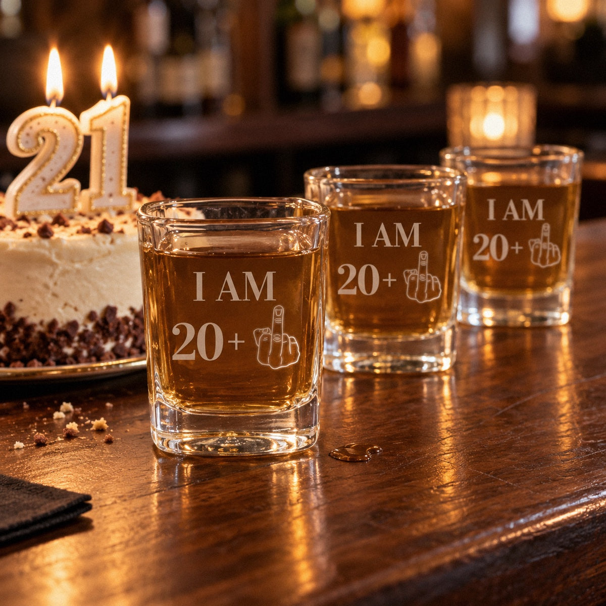 Three 21st birthday shot glasses engraved I AM 20 plus lined up on a bar beside a birthday cake with lit 21 candles