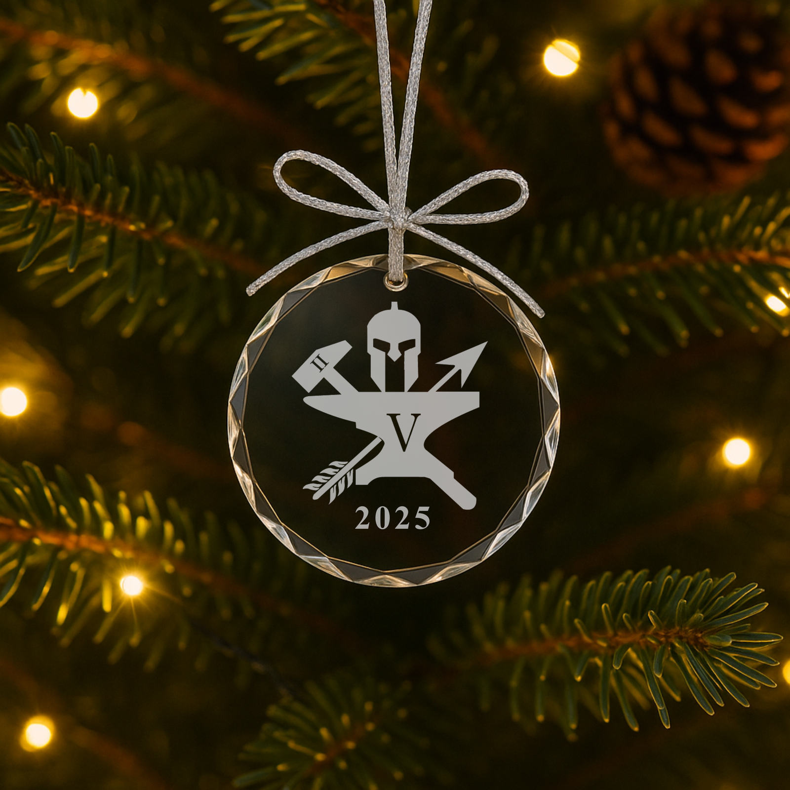 Round glass ornament with faceted border, engraved with sample tactical-themed logo featuring helmet, arrows, and "V" initial with 2025 date, hung from silver ribbon bow on evergreen branch with warm lights