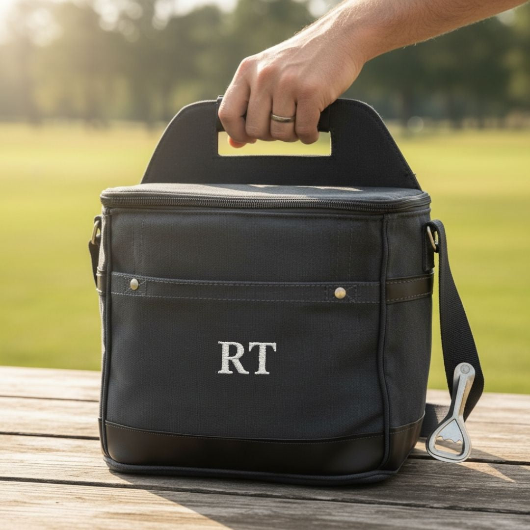 Black insulated cooler bag with monogrammed sample initials "RT" held by handle over outdoor setting with green field background