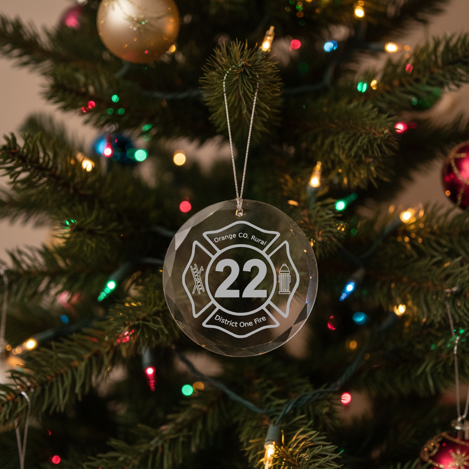 Round faceted glass ornament engraved with sample fire department logo reading "Orange CO. Rural District One Fire" and number 22, hanging from silver cord on illuminated Christmas tree.