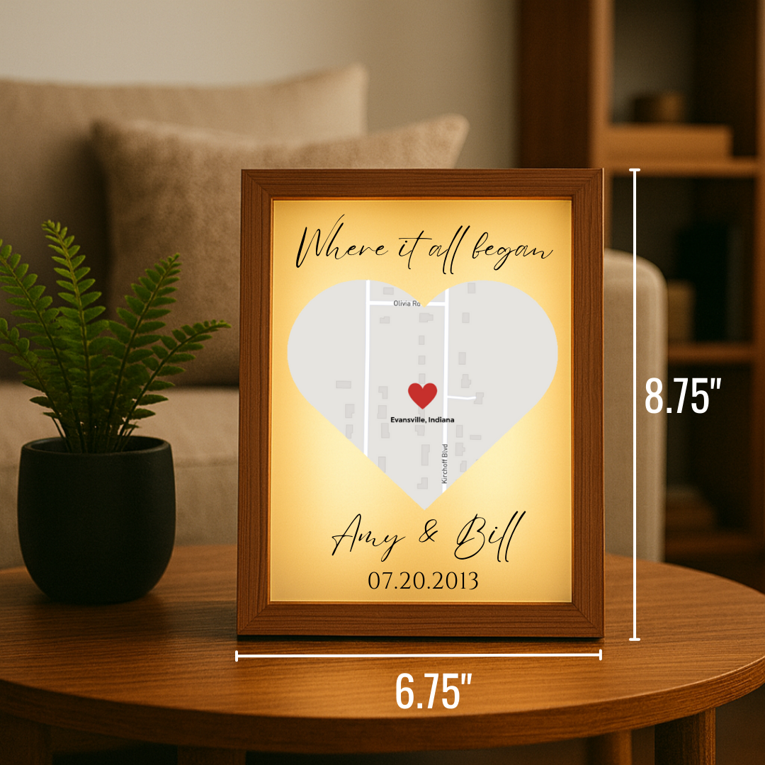 Framed light-up map art with sample personalization showing "Where it all began" text, heart marker on Evansville Indiana map, and names "Amy & Bell" with date 07.20.2013, displayed on wooden table with plant