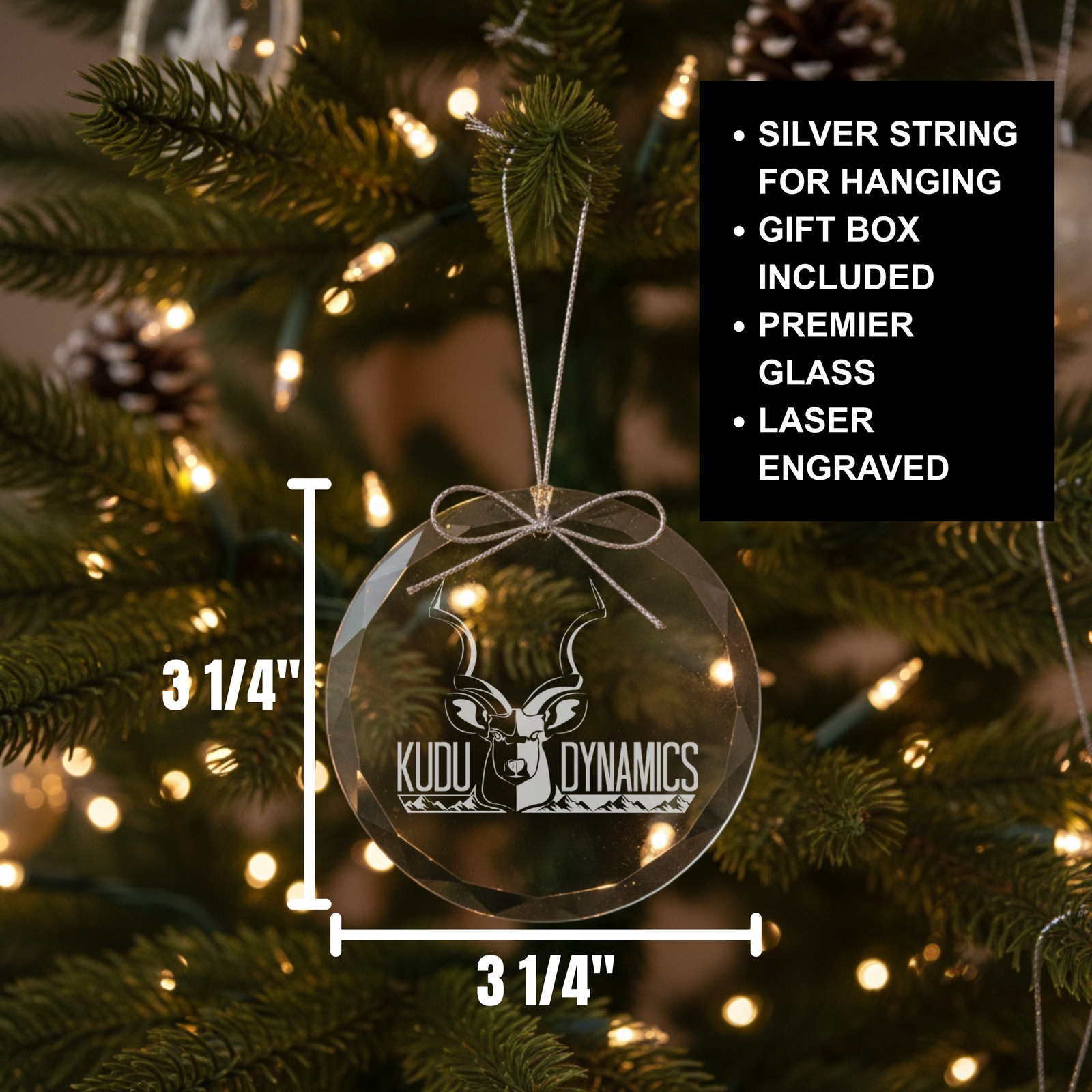 Round glass ornament with silver string hanging loop, laser-engraved with sample Kudu Dynamics logo design, displayed on evergreen branches with warm string lights