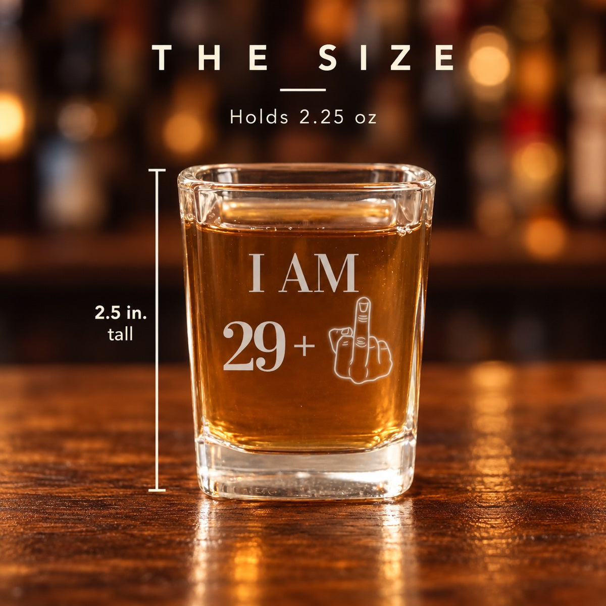 Size of the 30th shot glass engraved I AM 29 plus a middle finger: 2.5 inches tall and holds 2.25 ounces, shown on a walnut bar