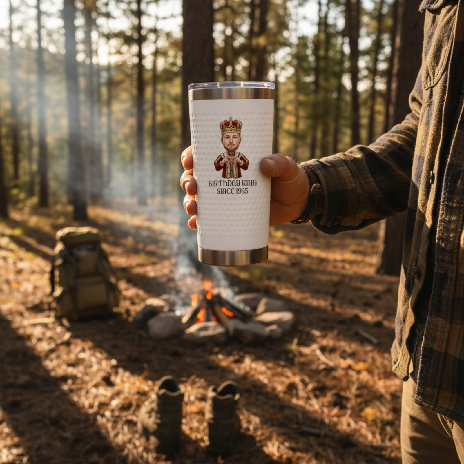 Insulated tumbler with stainless steel base held in hand, featuring sample design with cartoon king illustration and text "BIRTHDAY KING SINCE 1965" on white wrap, campfire scene background