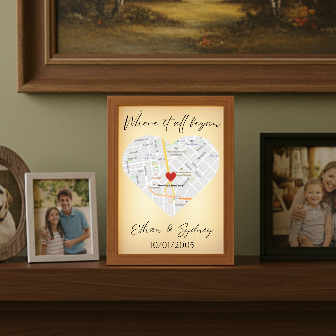 Framed map art with heart marker on New York City, personalized with sample names "Ethan & Sydney" and date "10/01/2005", illuminated by warm LED backlighting, displayed on wooden shelf with family photos