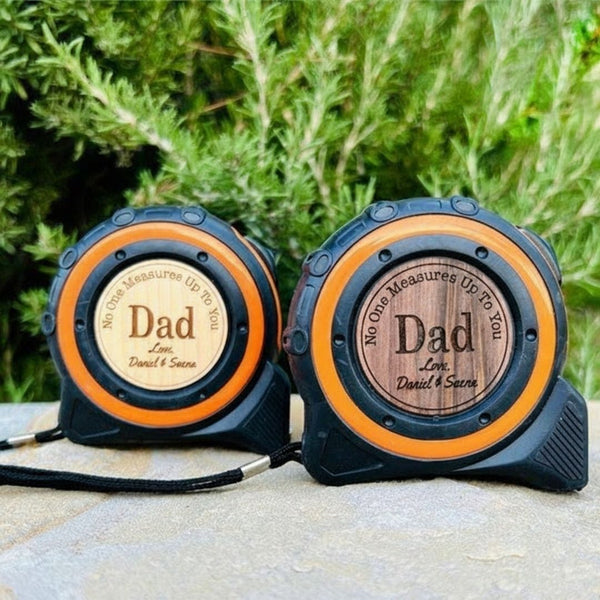 Two tape measures with blue casings and orange accents, each personalized with sample engraving reading "Dad" and "No One Measures Up To You" with names "Daniel & Saena