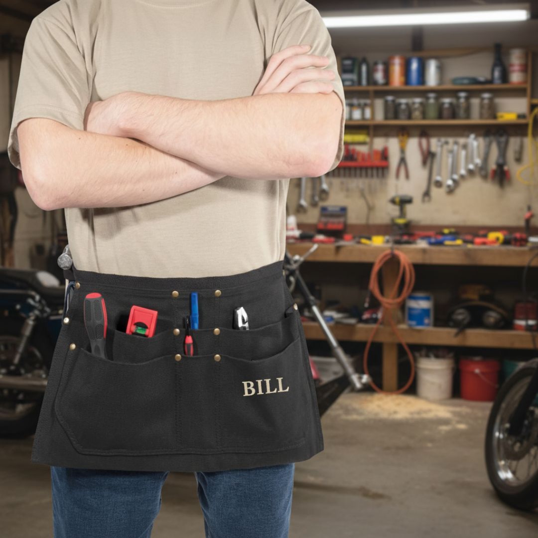 Black canvas tool belt with six pockets and hammer loop, personalized with sample name "BILL" embroidered in gold lettering, worn in workshop setting