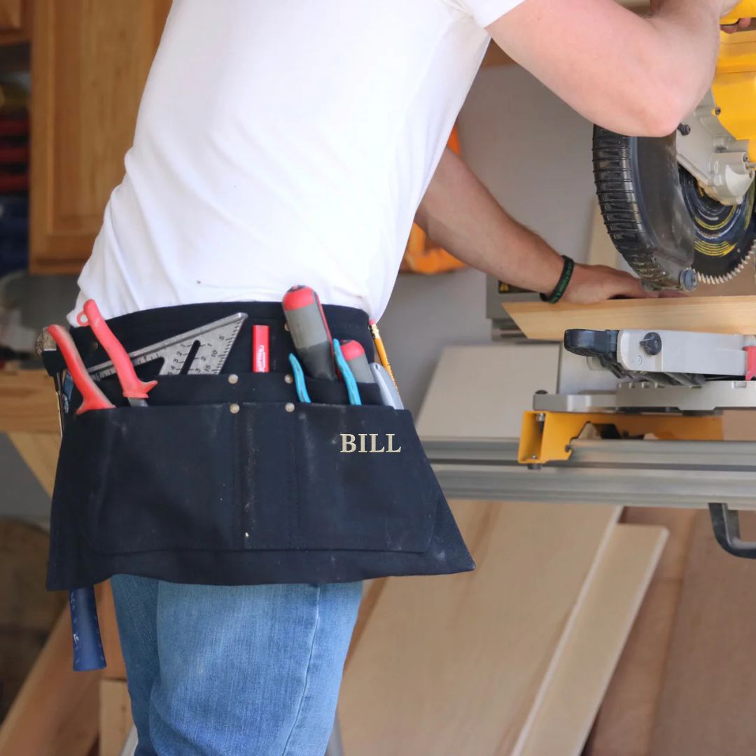 Navy canvas tool belt worn at waist, filled with hand tools and a level, personalized with sample name "BILL" in white lettering