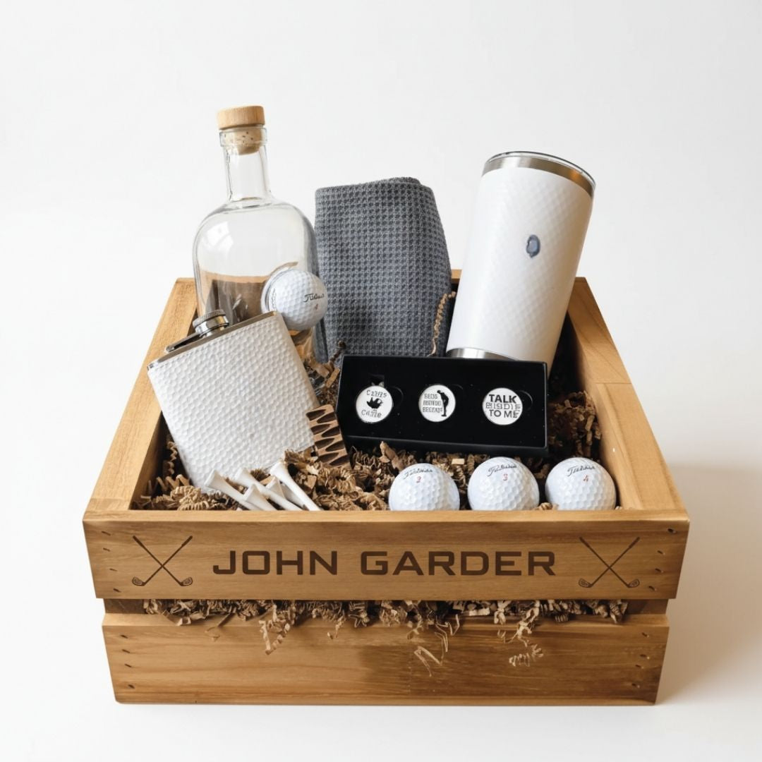 Wooden crate engraved with sample name "JOHN GARDER" and crossed clubs motifs, containing glass decanter with embedded golf ball, white insulated tumbler, golf ball markers in black box, gray and white waffle towels, and Titleist golf balls