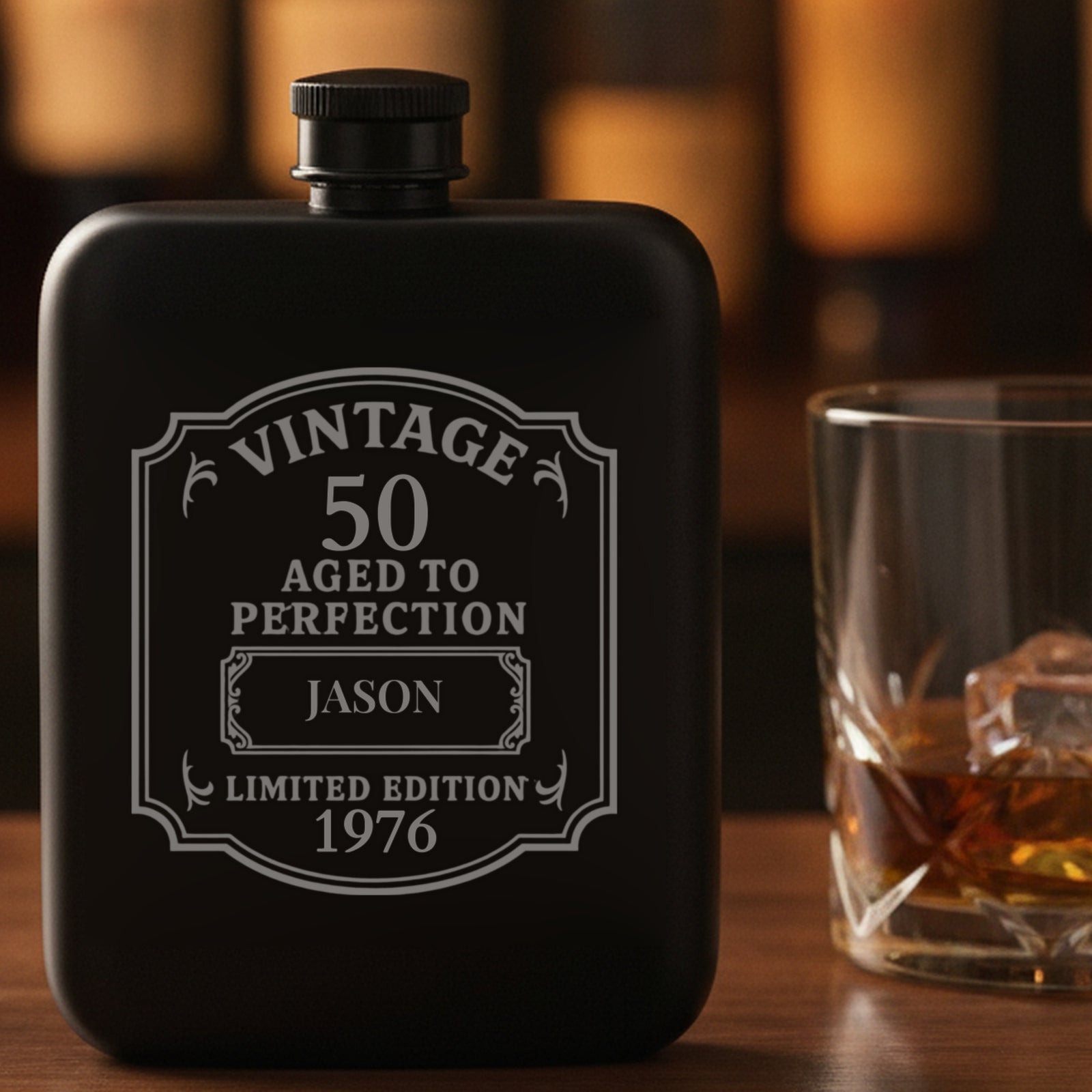 Engraved 50th birthday flask reading Vintage 50 Aged to Perfection, Jason, 1976, standing whole on a home bar beside a poured glass of bourbon.