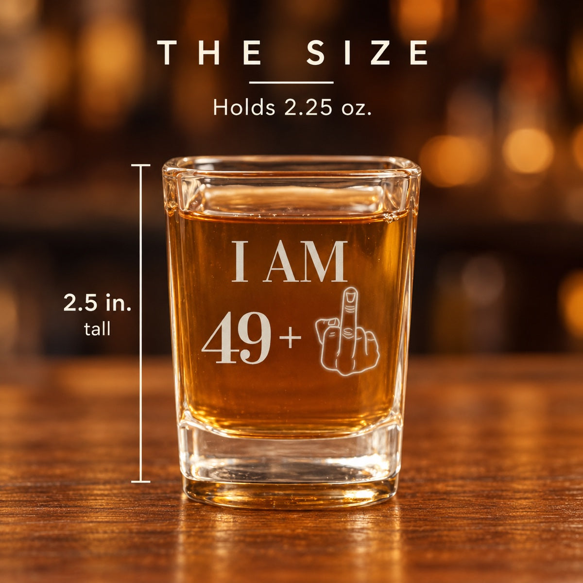 Size of the 50th shot glass engraved I AM 49 plus a middle finger: 2.5 inches tall and holds 2.25 ounces, shown on a walnut bar