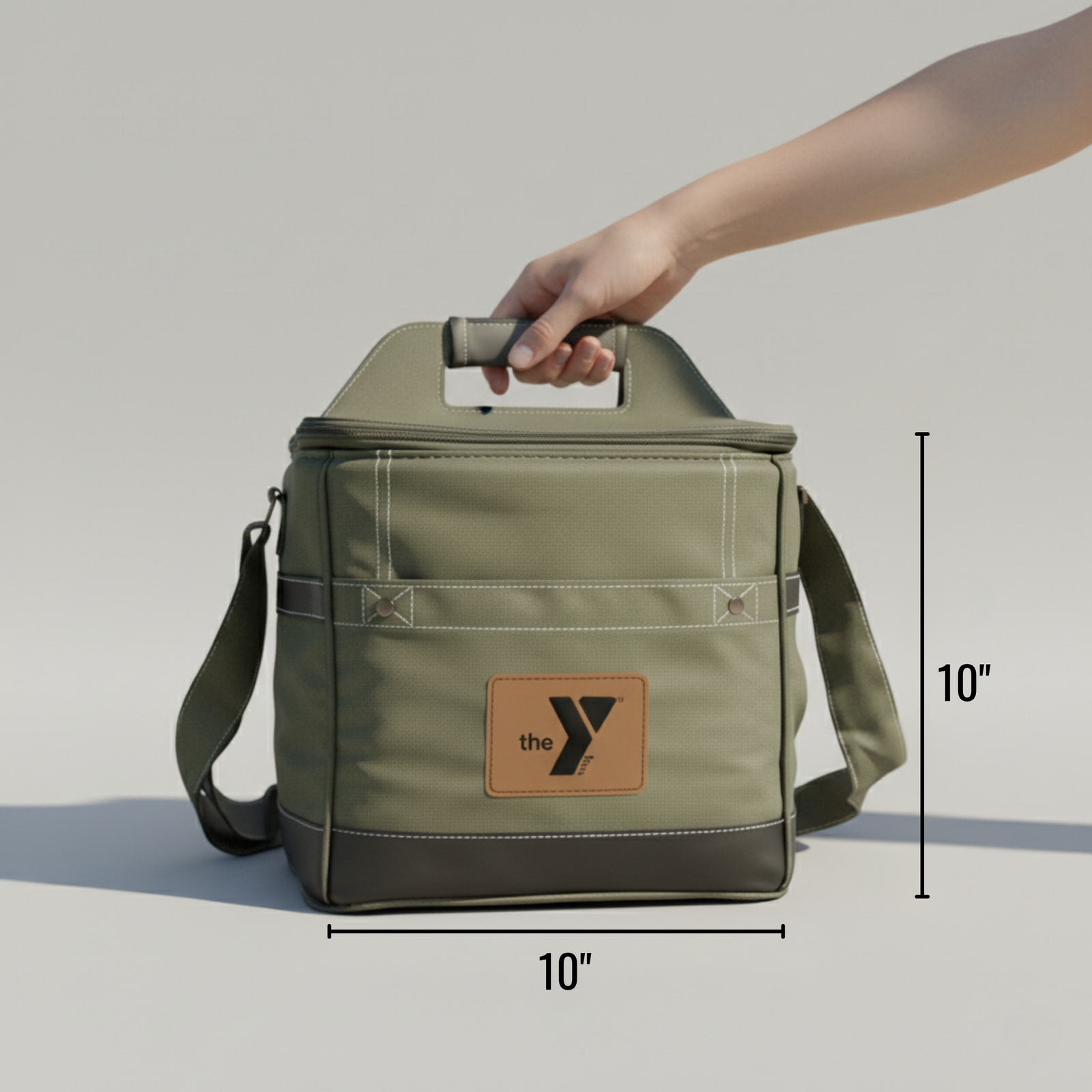 Sage-green insulated cooler bag with tan leather logo patch, shown held by carry handle, measuring 10 inches wide by 10 inches tall with shoulder strap