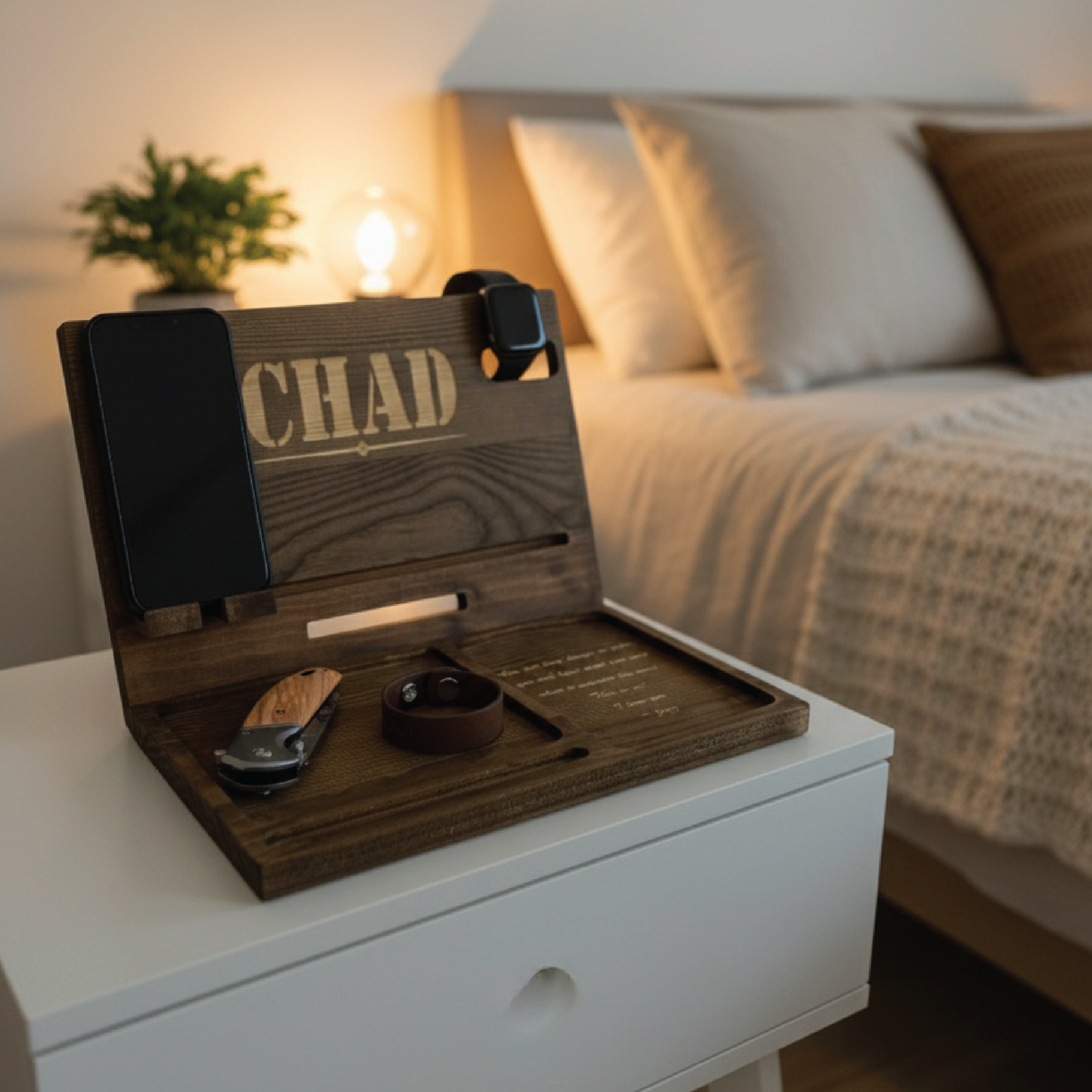 Wooden charging dock with sample engraving "CHAD" displayed on nightstand next to bed, holding smartwatch, phone, keys, and small folding knife
