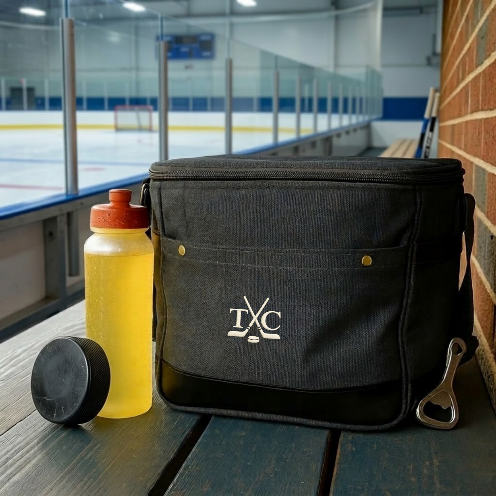 Black insulated cooler bag with embroidered hockey stick and puck logo, displayed on ice rink bench with yellow water bottle and hockey puck
