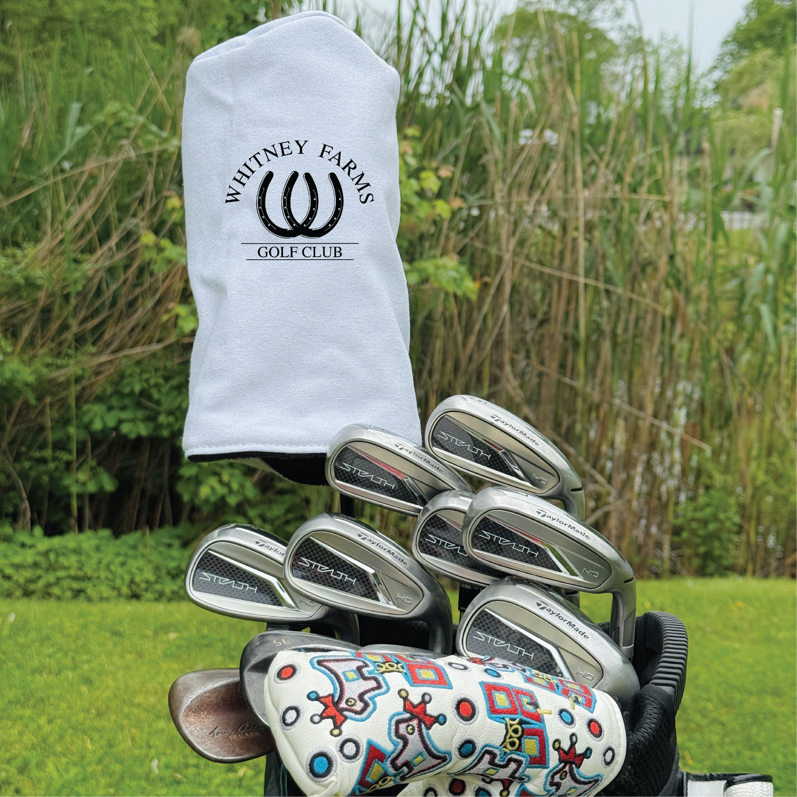 White golf club headcover with "Whitney Farms Golf Club" logo and horseshoe design displayed on bag with irons and colorful putter headcover outdoors