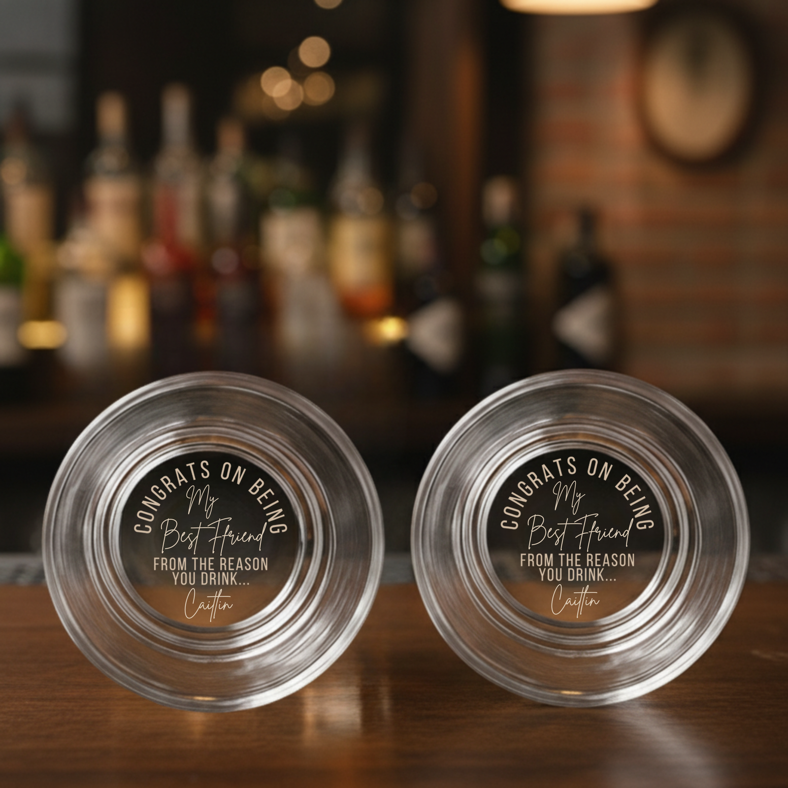 Pair of clear whiskey glasses with laser-etched sample personalization reading "Congrats on being my best friend from the reason you drink... Caitlin" on bottom, displayed on wooden bar counter with blurred bottles background