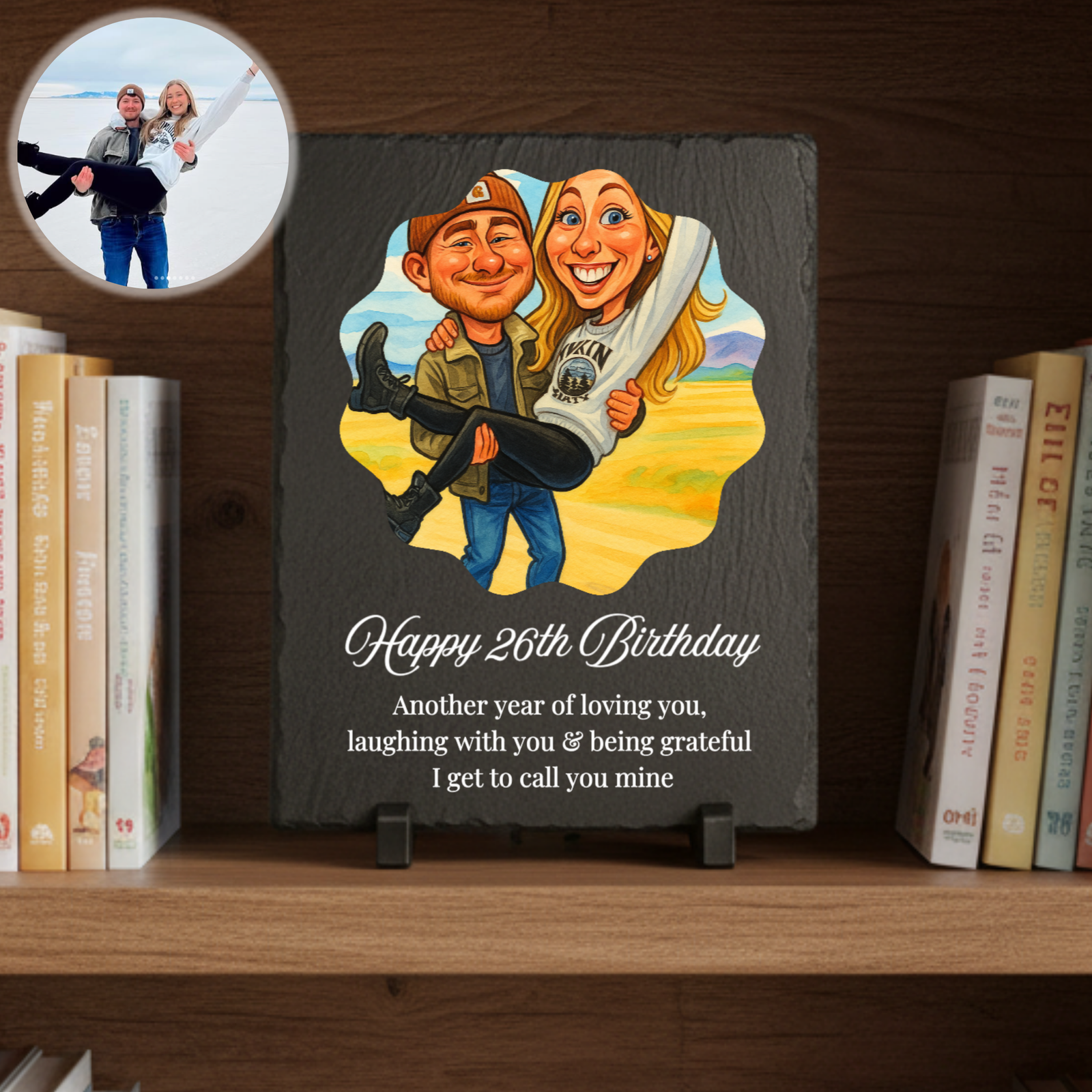 Slate plaque displayed on wooden shelf with books, featuring custom caricature of two people and sample text "Happy 26th Birthday" with personalized message below.