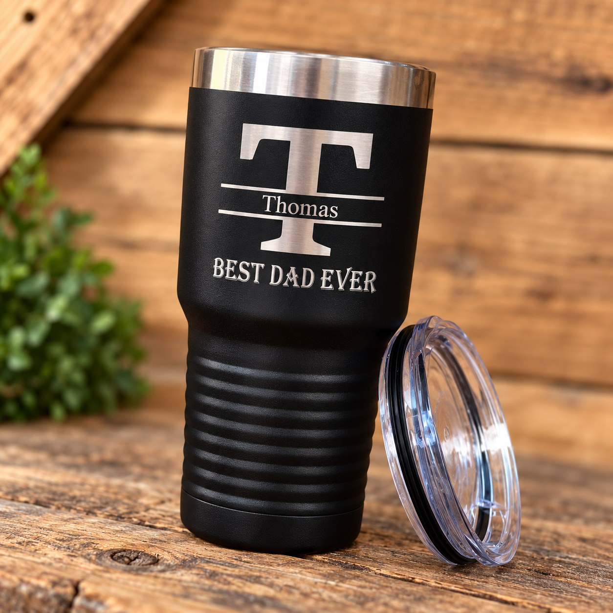 Black stainless steel tumbler with engraved monogram "T", name "Thomas", and text "BEST DAD EVER" shown with removable clear lid on wooden surface.
