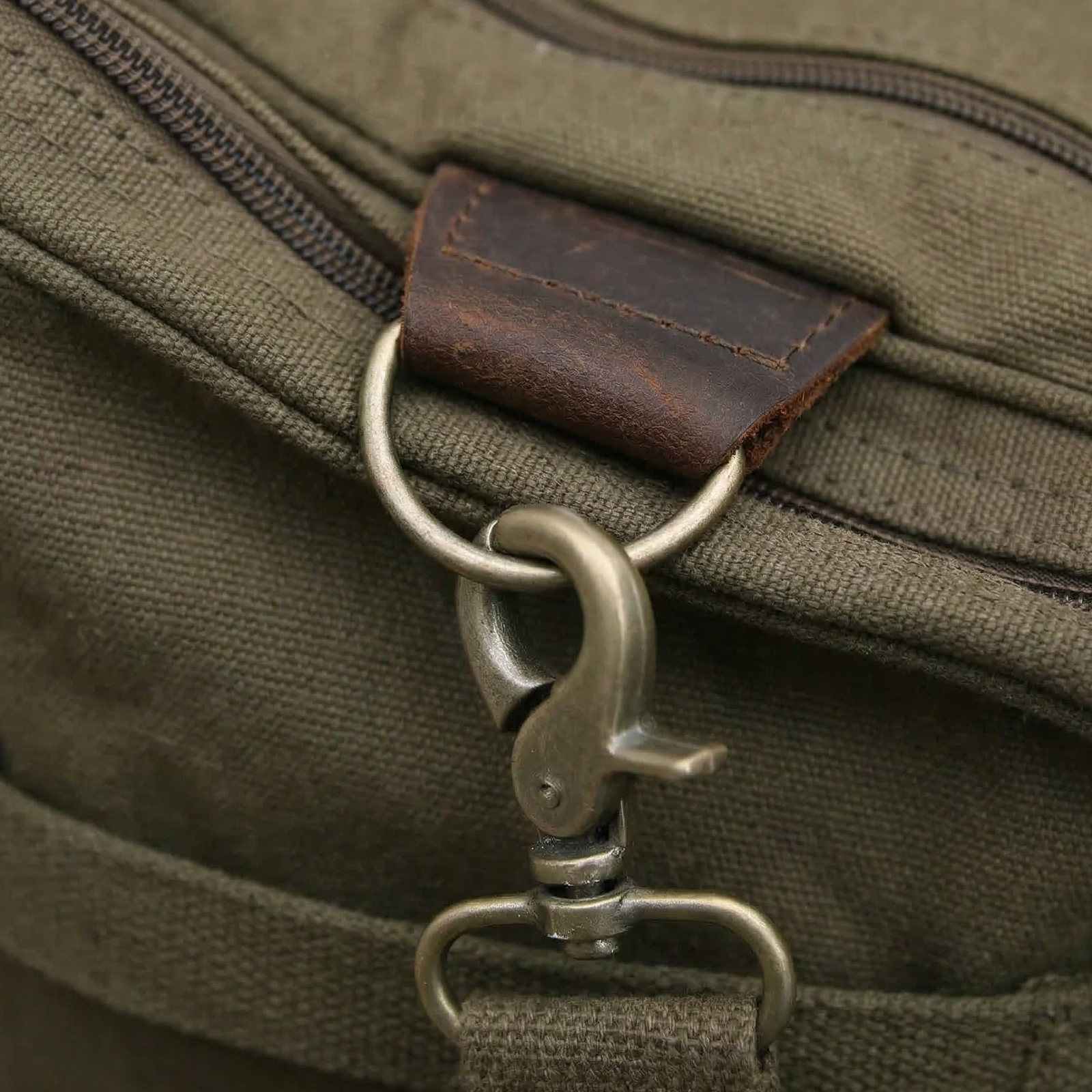 Canvas weekender bag in olive green with dark brown leather accent patch and metal snap hook detail attached to zippered pocket.