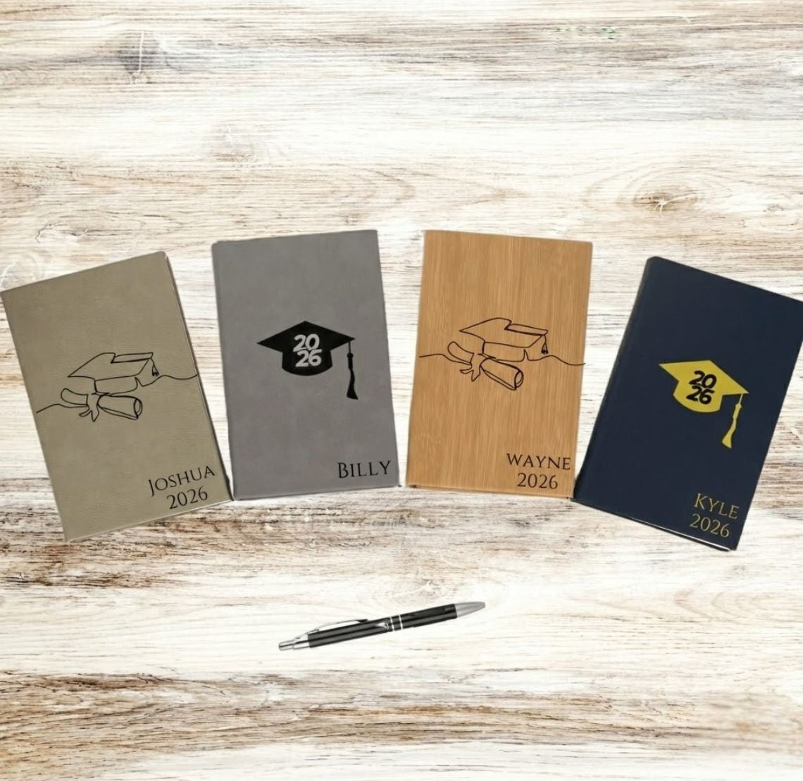 Four personalized notebooks with graduation-themed designs on a wooden surface, accompanied by a pen.