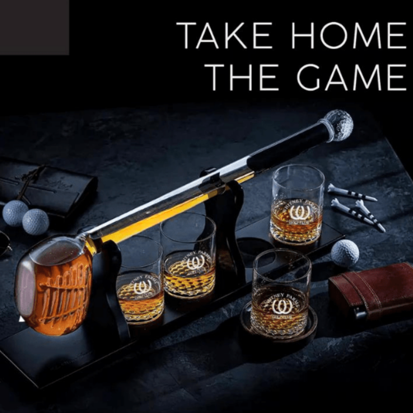 Golf club decanter set displayed with four rocks glasses containing sample logo engravings, wooden mug, baseball bat, golf balls, and leather accessories on dark surface