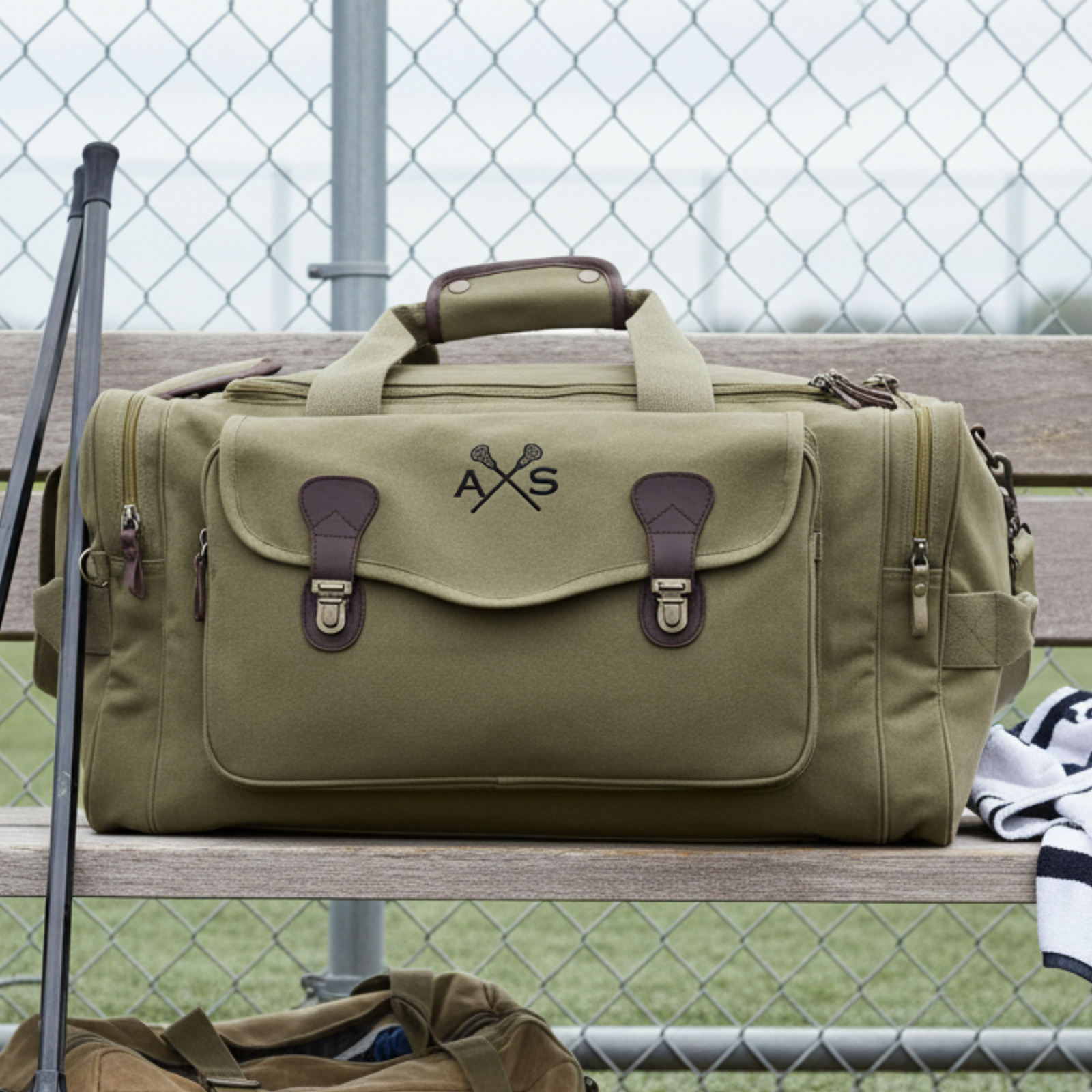 Tan canvas duffle bag with crossed lacrosse sticks and monogram "AXS" displayed on bleachers at a lacrosse field, brown leather handles and straps visible