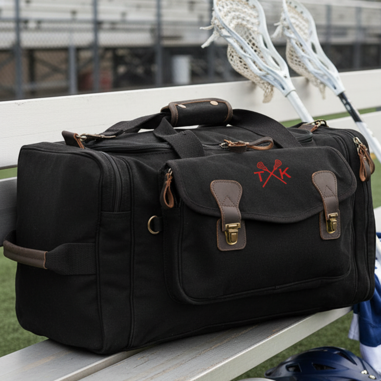 Black canvas lacrosse duffle bag with brown leather accents, dual brass buckles, and sample embroidered crossed sticks design in red on front panel, displayed on field bench with lacrosse sticks behind