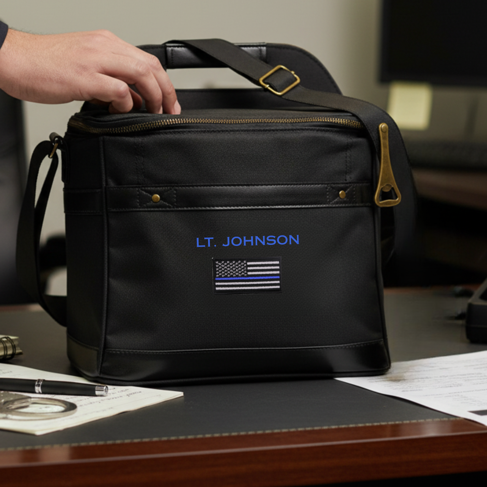 Black cooler bag with sample personalization reading "LT. JOHNSON" and thin blue line flag embroidered on front, brass hardware and carry handles, sitting on desk.