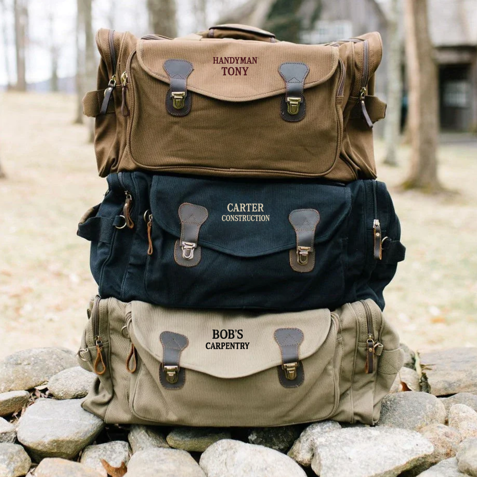 Three personalized canvas duffle bags stacked vertically, showing sample engravings reading "HANDYMAN TONY," "CARTER CONSTRUCTION," and "BOB'S CARPENTRY" in tan, navy, and khaki colors