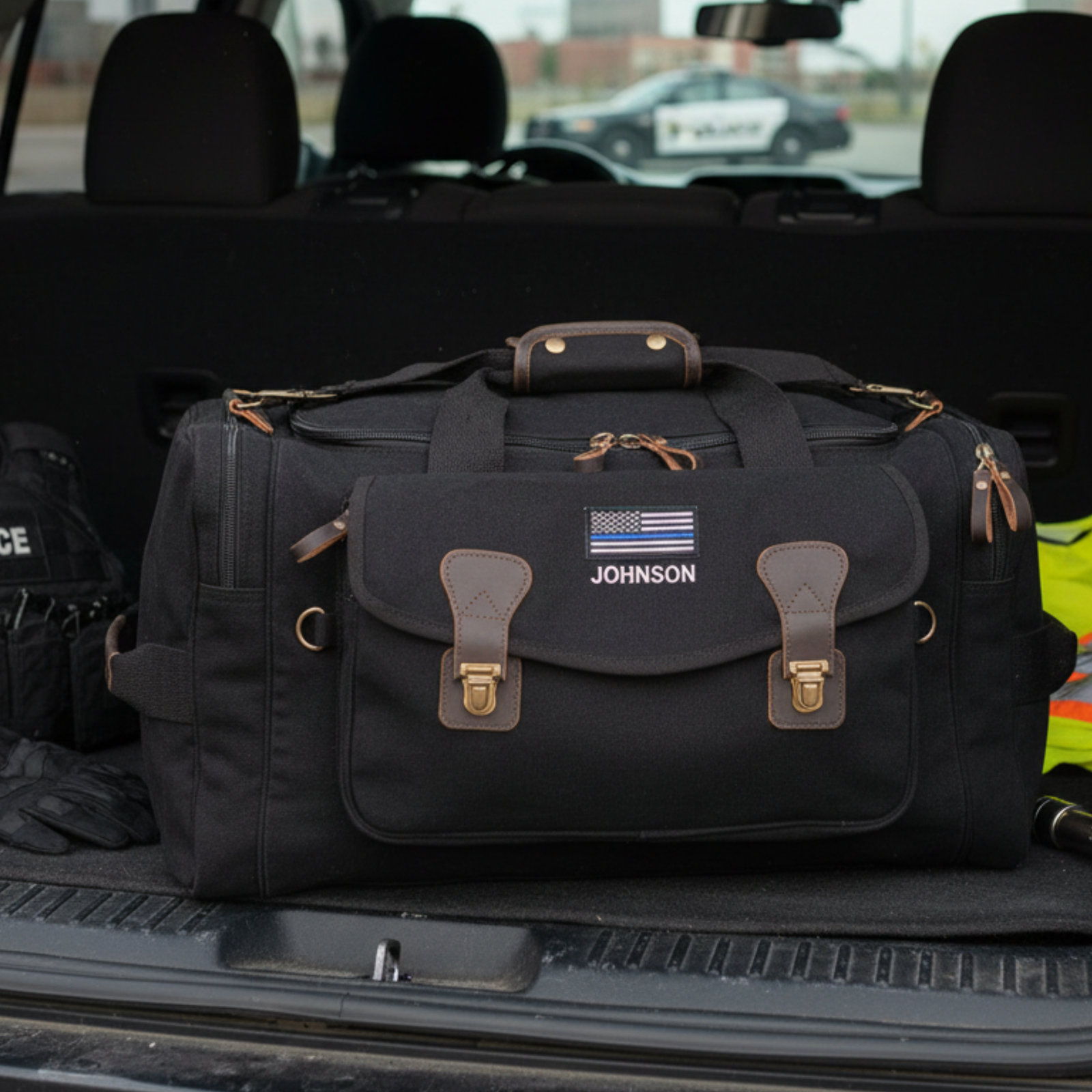 Black canvas duffle bag with brown leather handles and accents, personalized with sample name "JOHNSON" and thin blue line flag emblem, positioned in vehicle trunk.