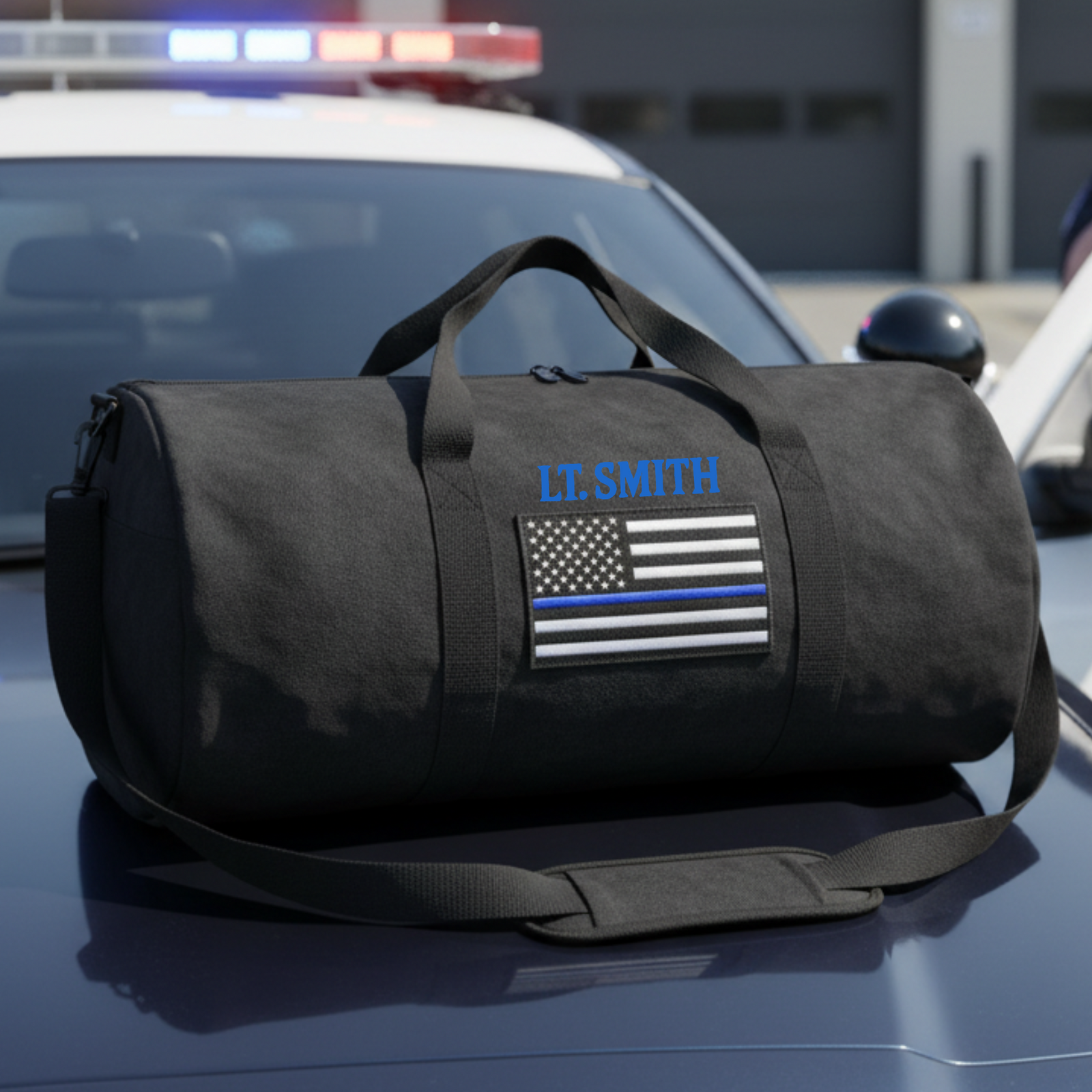 Black canvas duffle bag with shoulder strap, personalized with sample name "LT. SMITH" in blue embroidery and thin blue line flag patch on front.