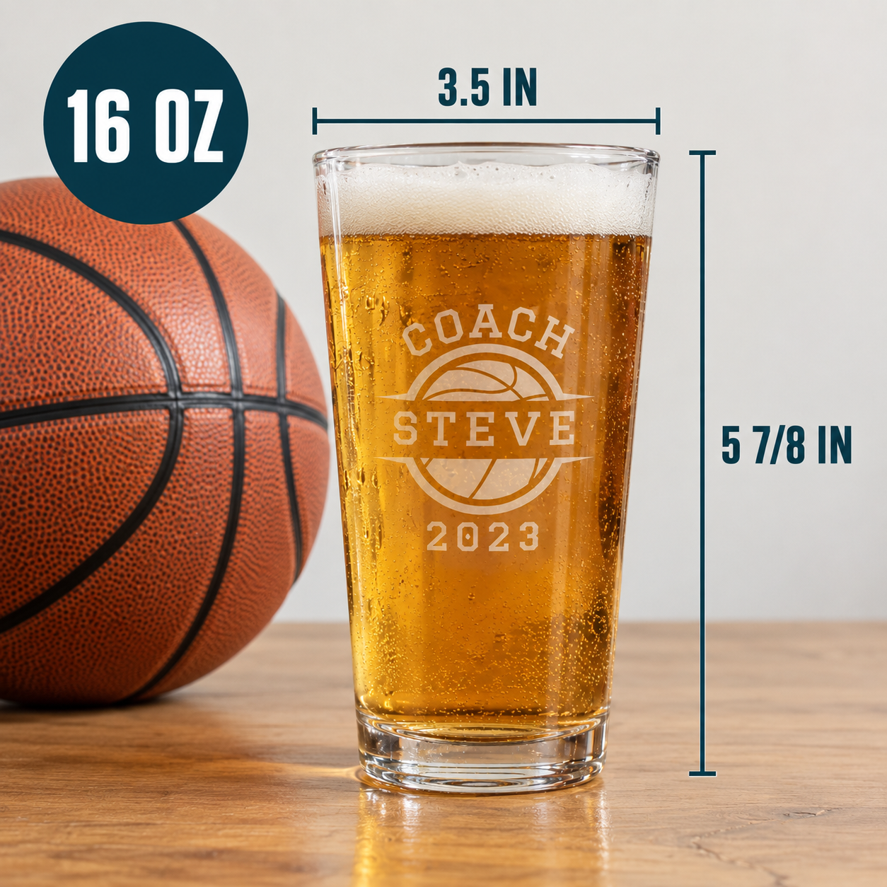 16 oz pint glass filled with beer, engraved with "COACH STEVE" and basketball design with year "2023", displayed next to basketball