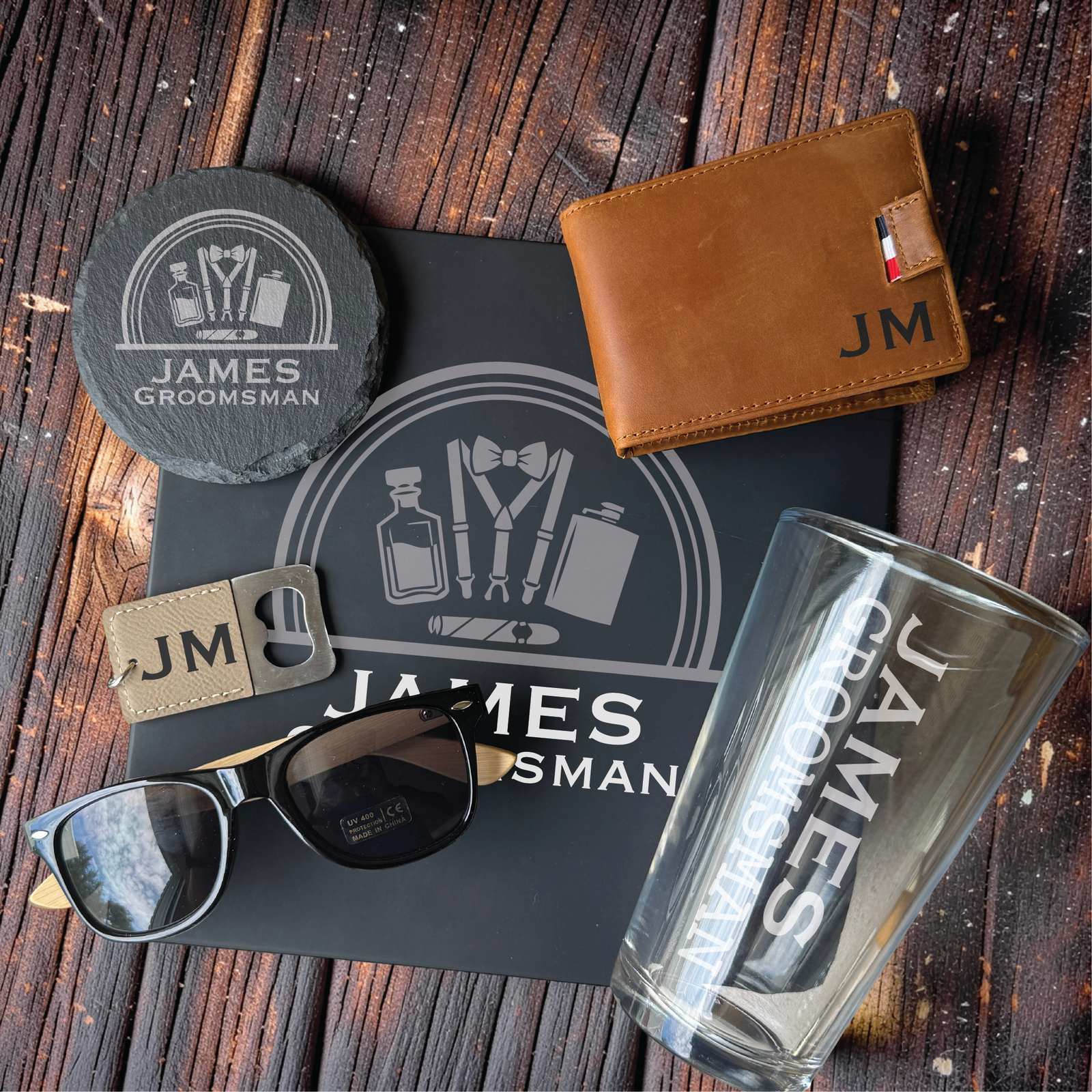 Flat lay of a customized men's gift set with engraved slate coaster, tan leather wallet, bottle opener keychain, polarized sunglasses, and a monogrammed pint glass on rustic wood.