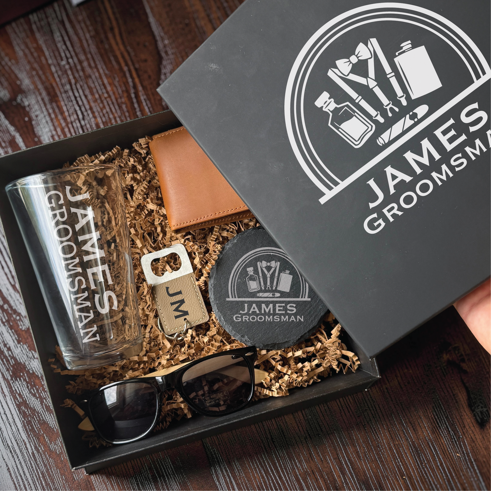 Personalized gift box for men with an engraved lid, holding a customized pint glass, leather wallet, coaster, sunglasses, and JM bottle opener keychain.