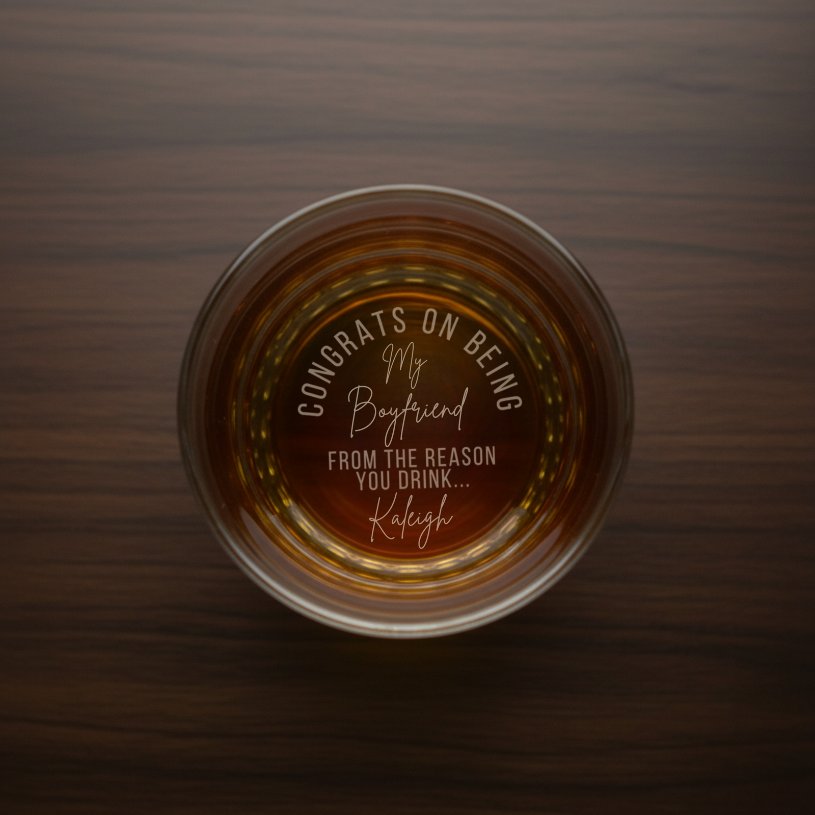 Overhead view of whiskey glass filled with amber liquid, laser-engraved bottom reads "Congrats on being my boyfriend from the reason you drink Kayleigh