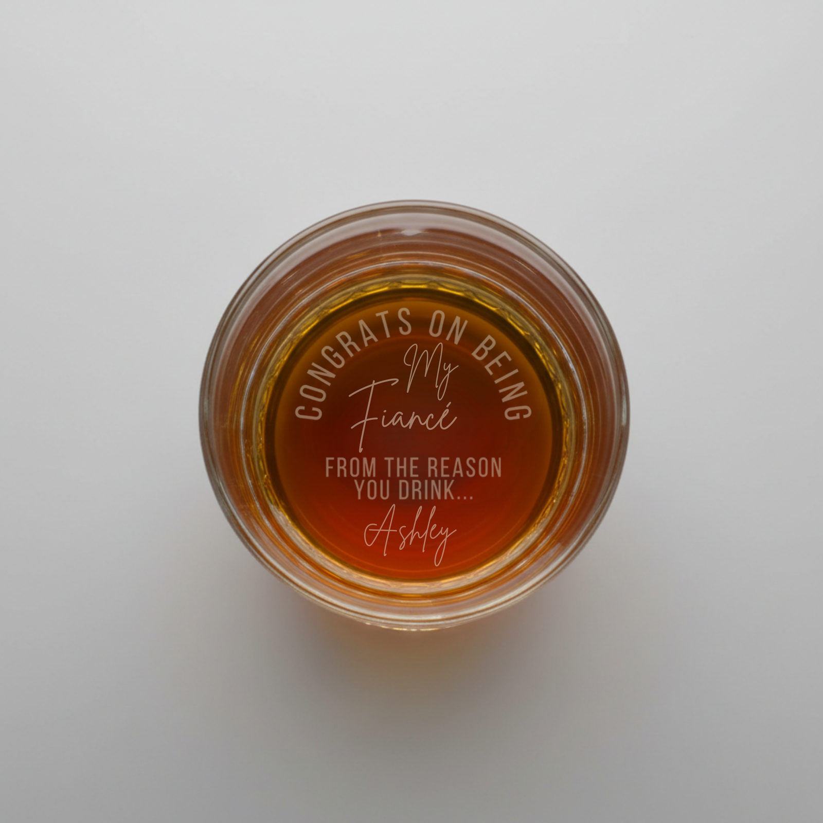 Overhead view of whiskey glass with sample personalization laser-etched on bottom reading "Congrats on being my Fiancé from the reason you drink... Ashley