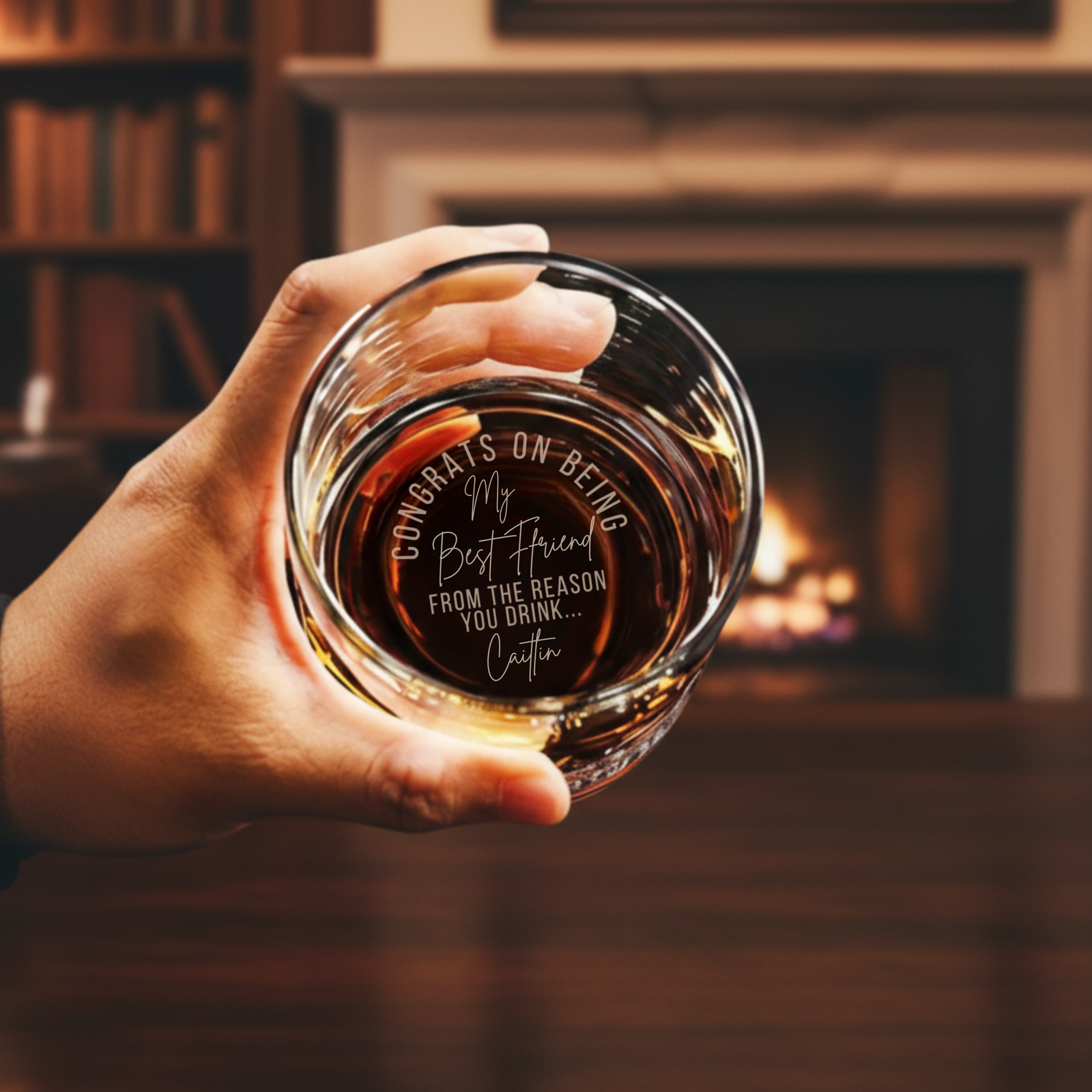 Hand holding whiskey glass filled with amber liquid, displaying laser-etched sample message "Congrats on being my Best Friend from the reason you drink... Caitlin" on bottom, fireplace blurred in background