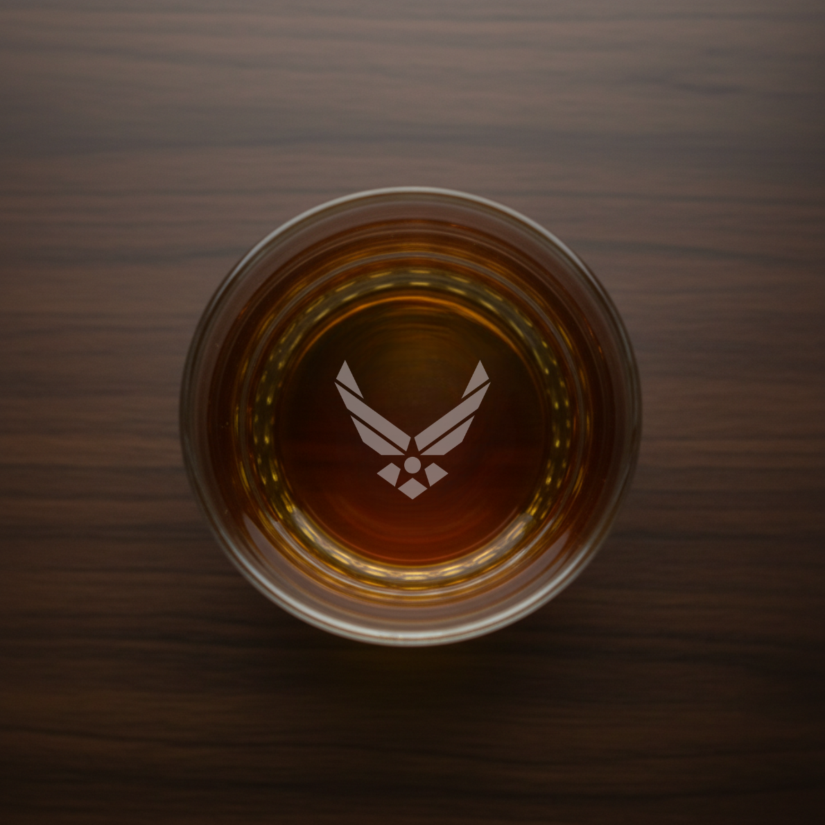 Whiskey glass with engraved Air Force wings symbol, filled with amber liquid, shot from above on wooden surface
